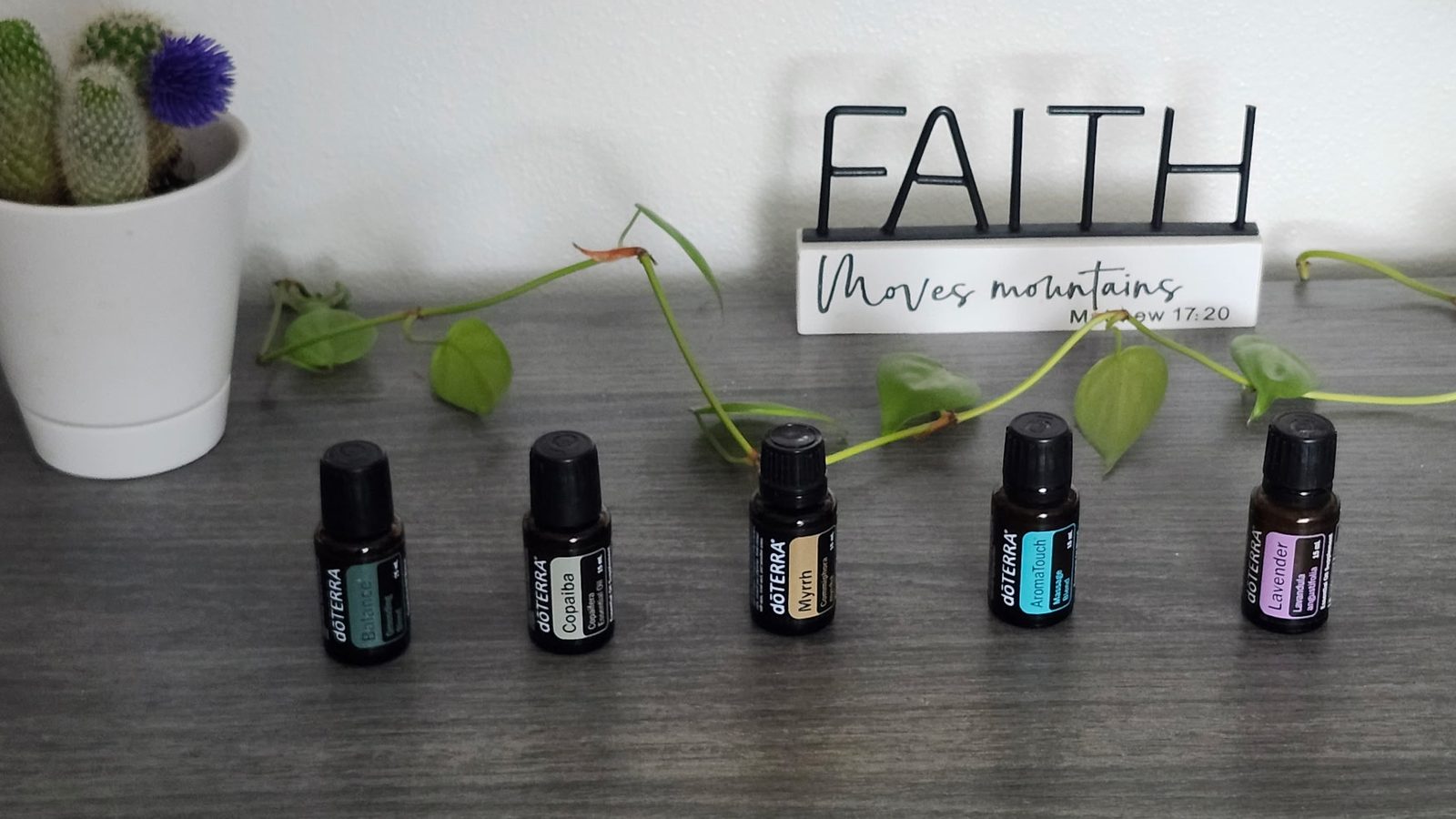 Five dōTERRA essential oil bottles (Balance, Copaiba, Myrrh, AromaTouch, and Lavender) on a wood surface, with a small cactus and trailing ivy in front of a sign reading 'Faith moves mountains — Matthew 17:20'.